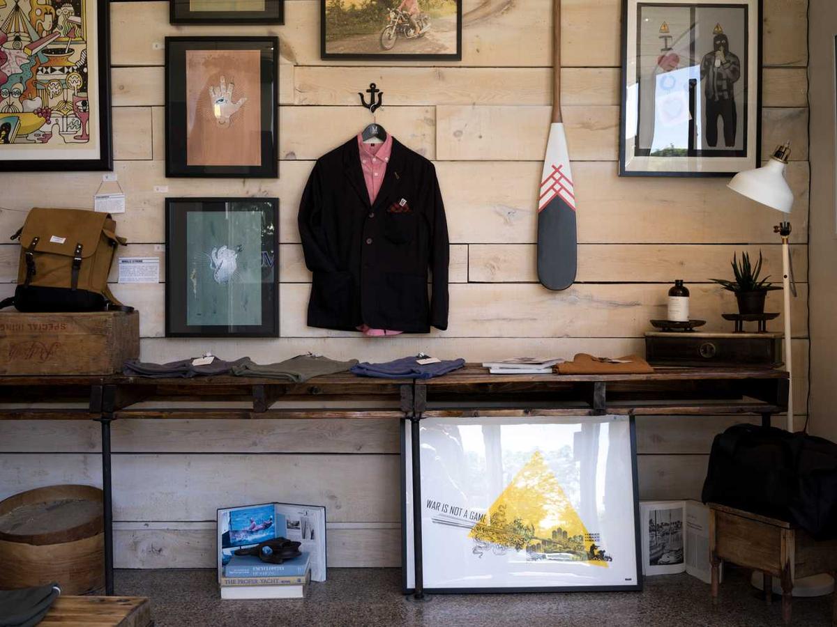 A wooden-walled boutique interior featuring framed artwork, a jacket on a hook, folded clothes on a table, and various decor items arranged neatly.