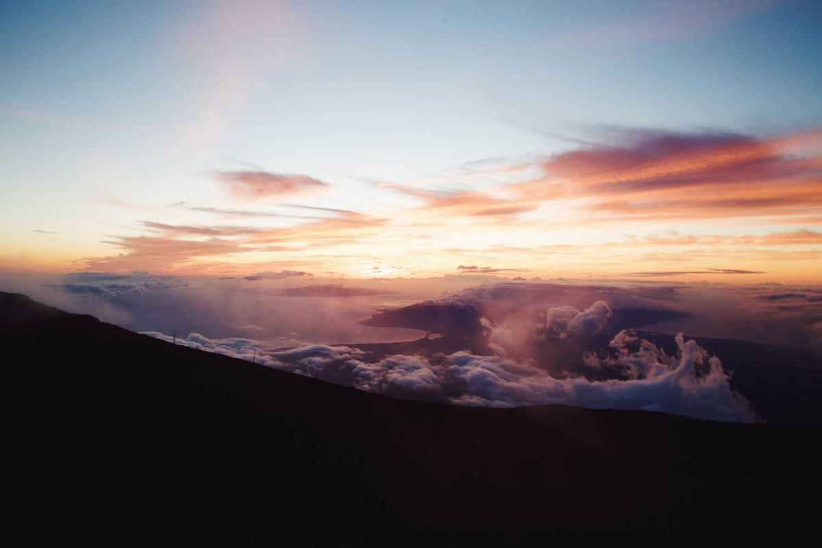 A wide scenic view of a colorful sunset sky with pink and orange clouds above a mountain ridge and thick cloud layers below.